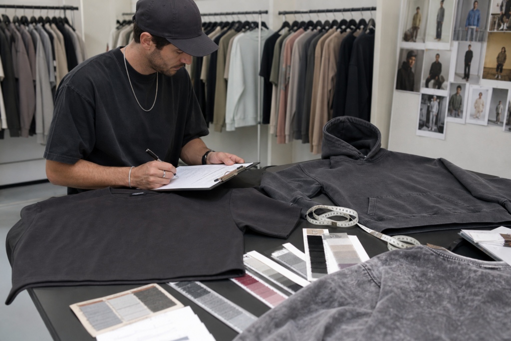 Apparel buyer in a showroom reviewing streetwear samples: a boxy heavyweight T-shirt and hoodie on a table with measurement tape, fabric swatches, and a washed reference sample, taking notes on a clipboard, realistic, no brands